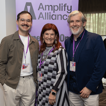 Three attendees posing together in front of an Amplify Alliance banner.